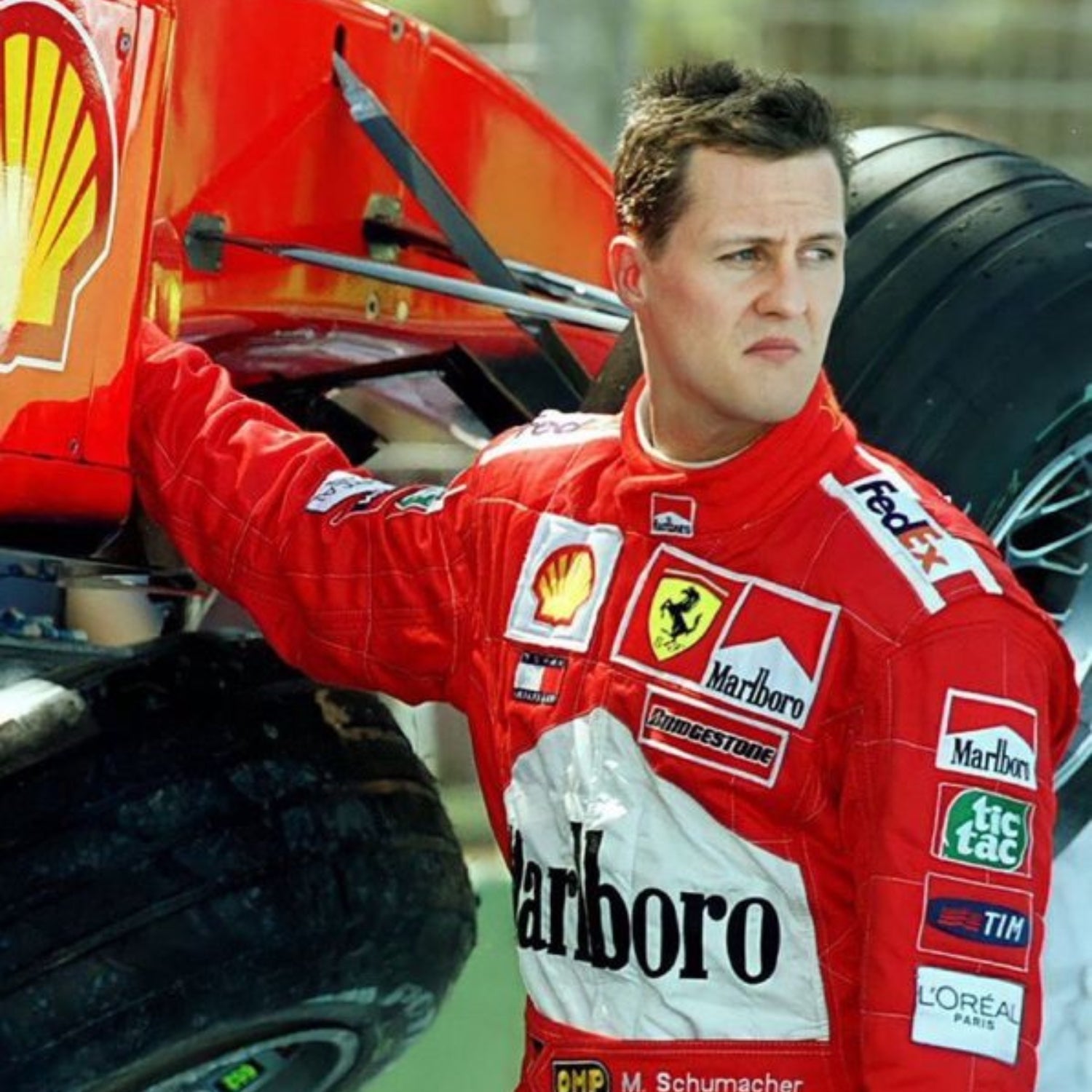 Michael Schumacher in a red Ferrari 2002 f1 racing suit with various sponsor logos next to a Formula 1 car.