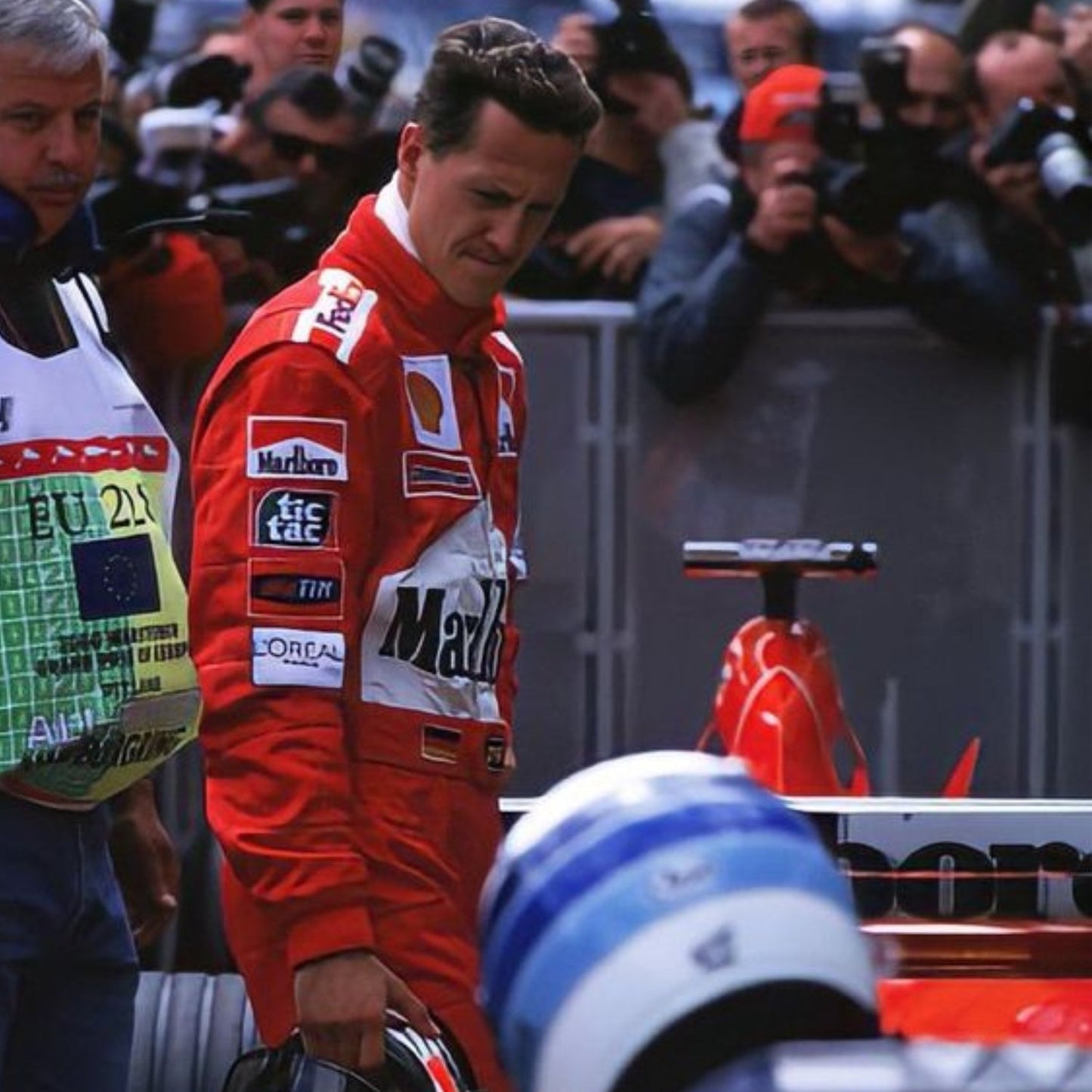 Michael Shumacher in a red racing suit with various sponsor logos, standing in front of a Formula 1 car.